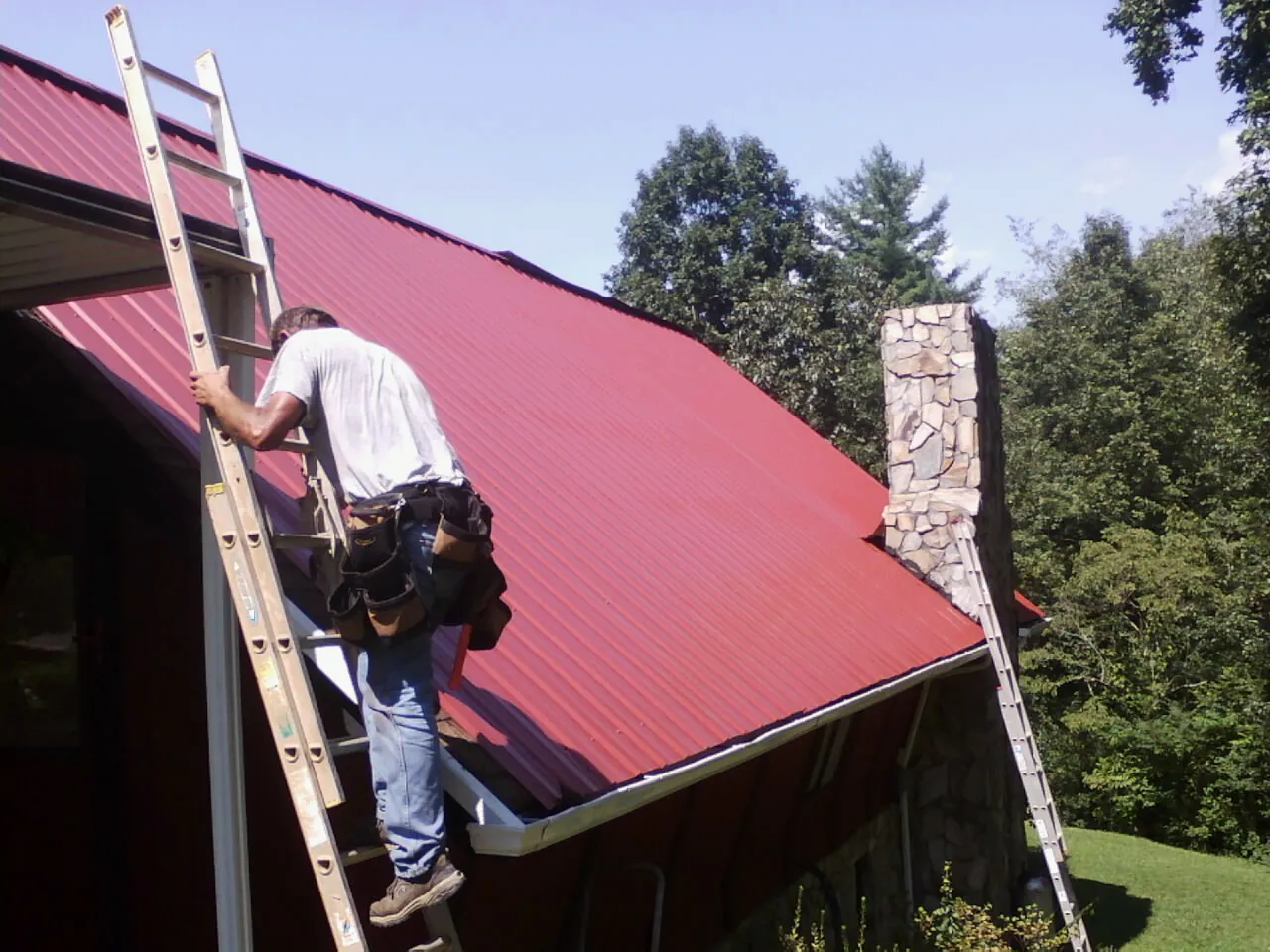 Roofer on a ladder beside a red metal roof and stone chimney — Soffit & Fascia Repair by Roofing Brothers in La Honda
